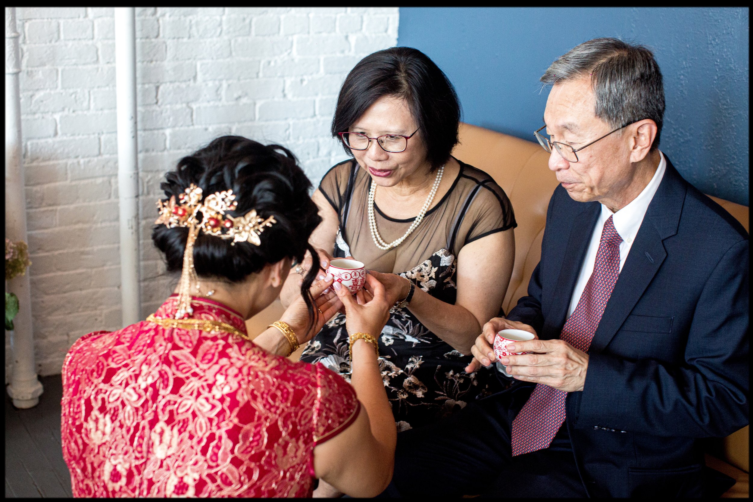 An Asian woman wearing glasses and a black lace top shares a tea cup with another woman dressed in traditional red Asian attire with gold jewelry. They are seated with an older Asian man in a suit, all holding small tea cups during a cultural tea ceremony in a modern setting.