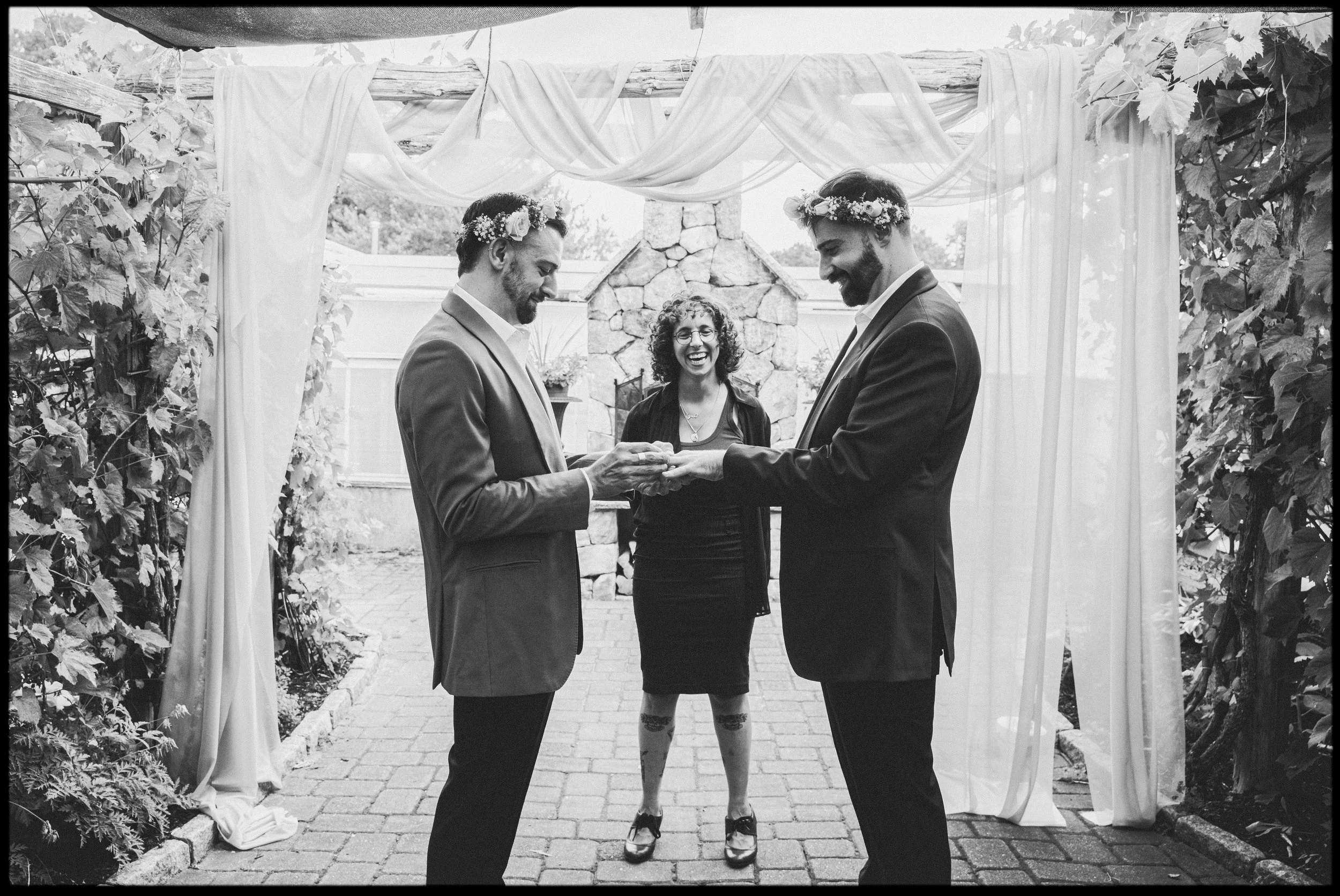 Two men in suits and flower crowns exchanging vows during a wedding ceremony outdoors, with a woman officiating, surrounded by a decorated outdoor setting.