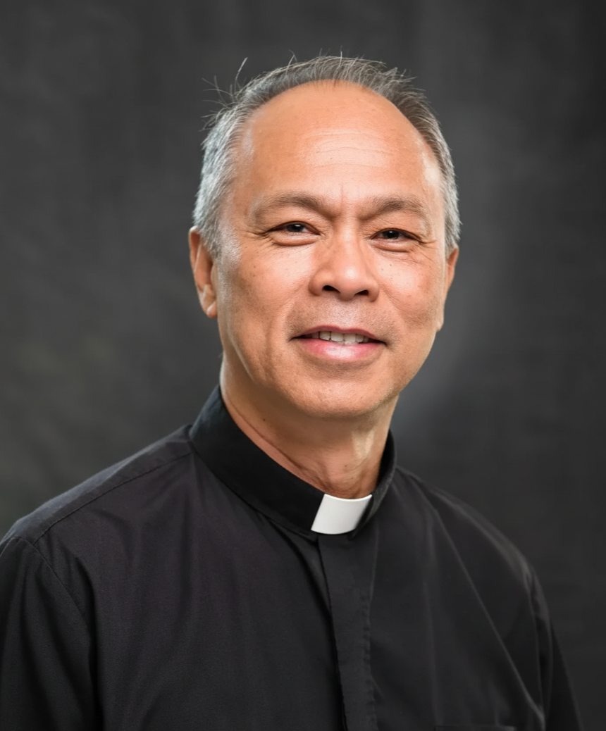 Portrait of a smiling male priest or clergyman wearing a black clerical shirt with a white clerical collar, standing against a dark background.