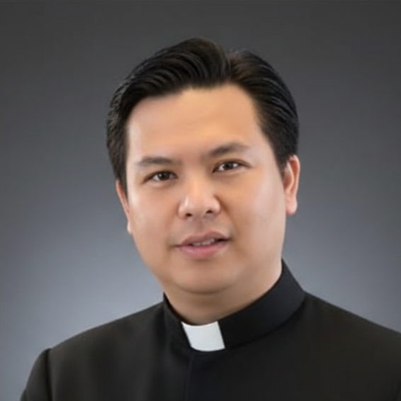 Portrait of a young male priest in a black clerical shirt with a white collar, standing against a gray background.