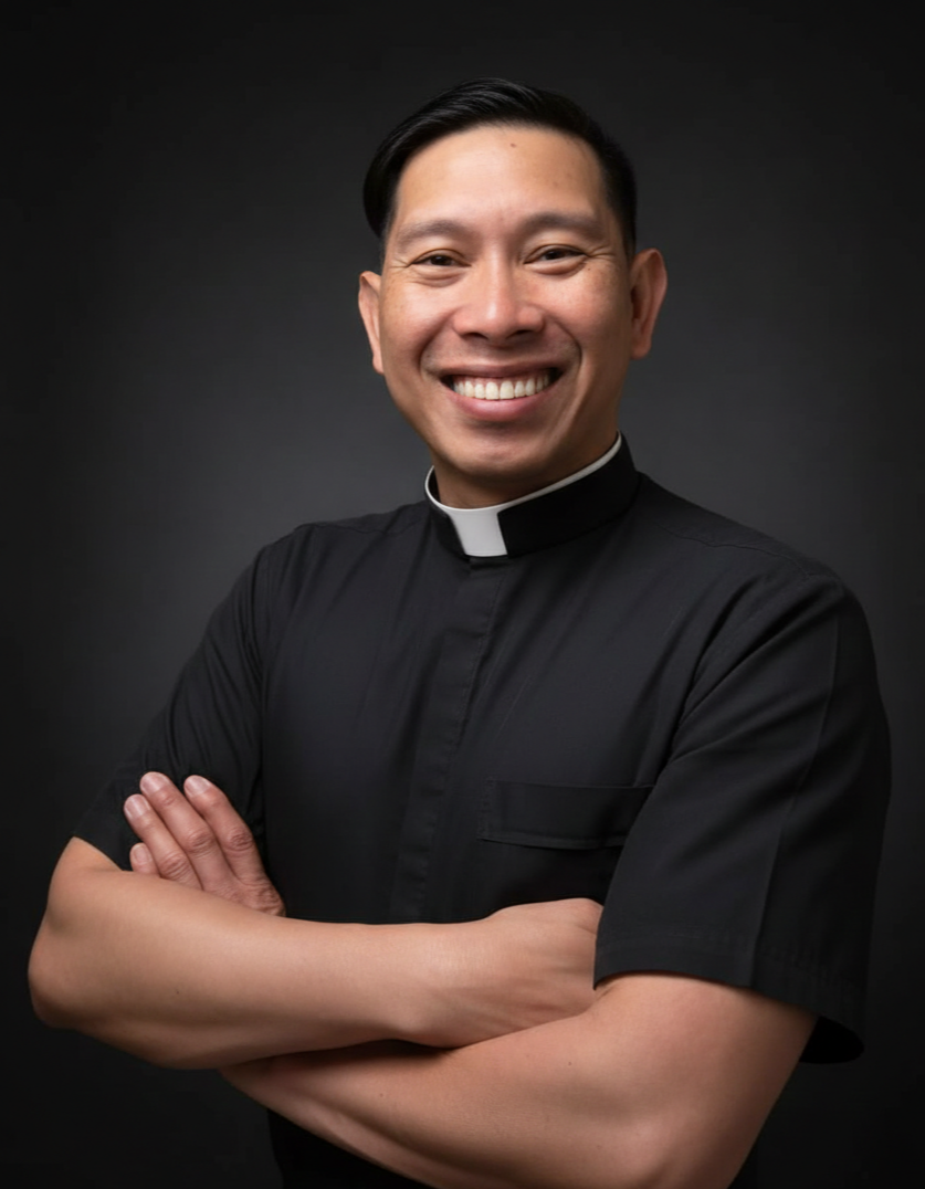 Smiling man wearing clergy attire with arms crossed, dark background.