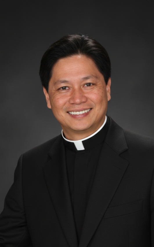 Portrait of a smiling priest wearing a black suit with a clerical collar against a dark grey background.