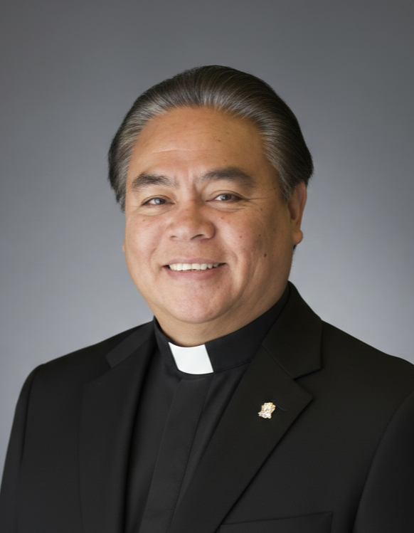 A smiling priest in a black suit with a clerical collar, against a neutral background.