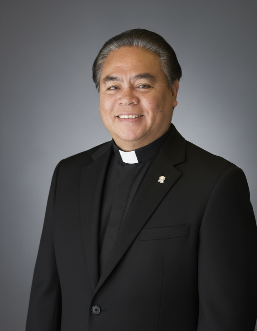 Portrait of a smiling man dressed as a Catholic priest in a black suit with a clerical collar, against a gray background.
