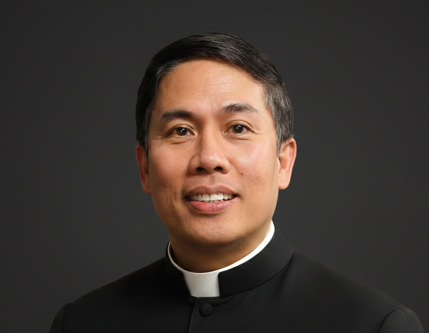 Portrait of a man in a black clerical shirt with a white clerical collar, against a dark background.