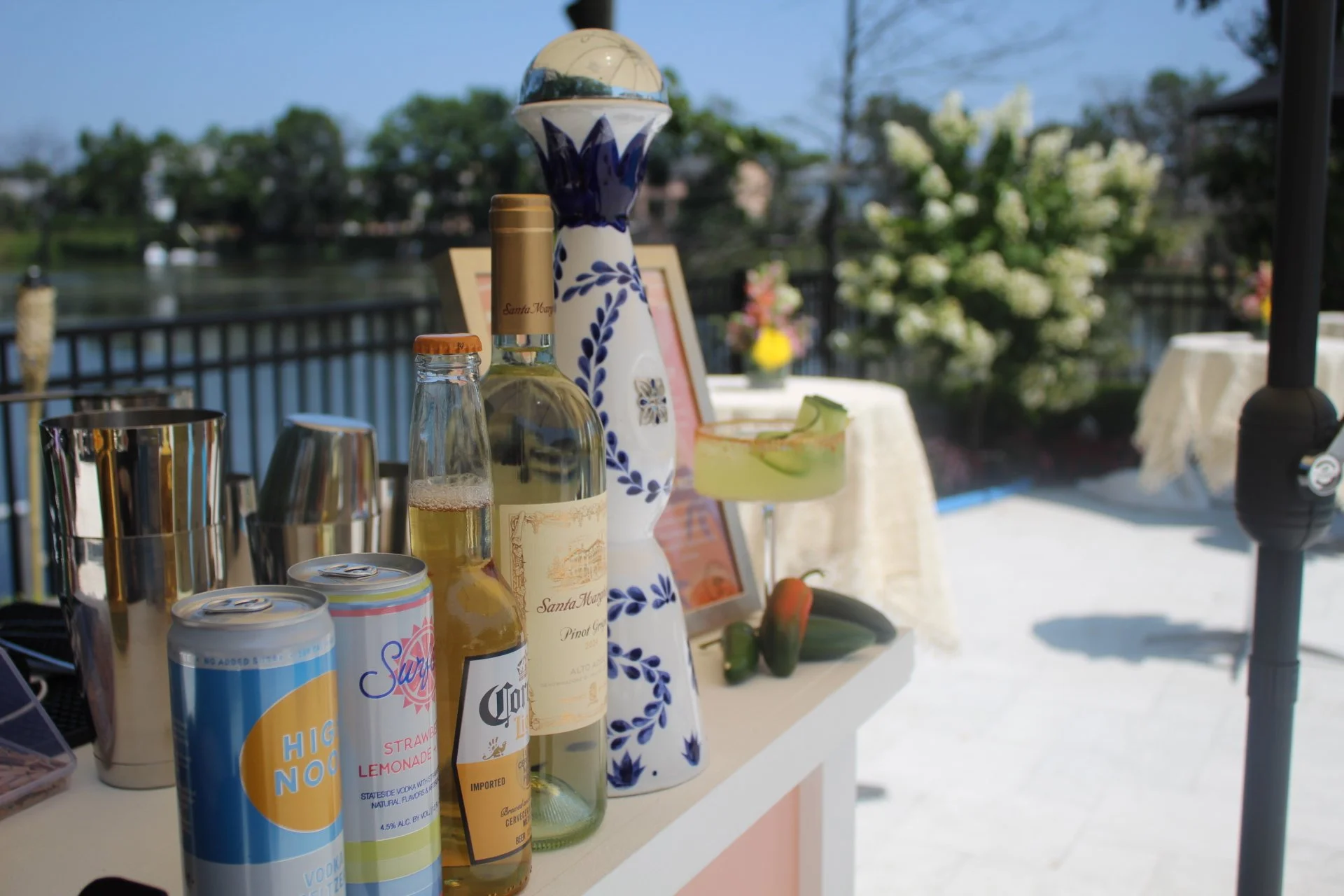 Outdoor bar setup with bottles of alcohol, cans of soda, cocktail shaker, and decorative ceramic vase, overlooking a body of water with trees and flowering plants in the background.