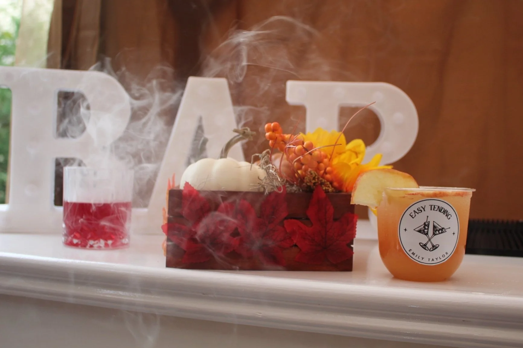 Decorative autumn-themed display with a small pumpkin, orange berries, yellow flower, and apple slices in a container, a red and white candle, and a drink with a lemon slice on a white surface with a large letter 'B' in the background.