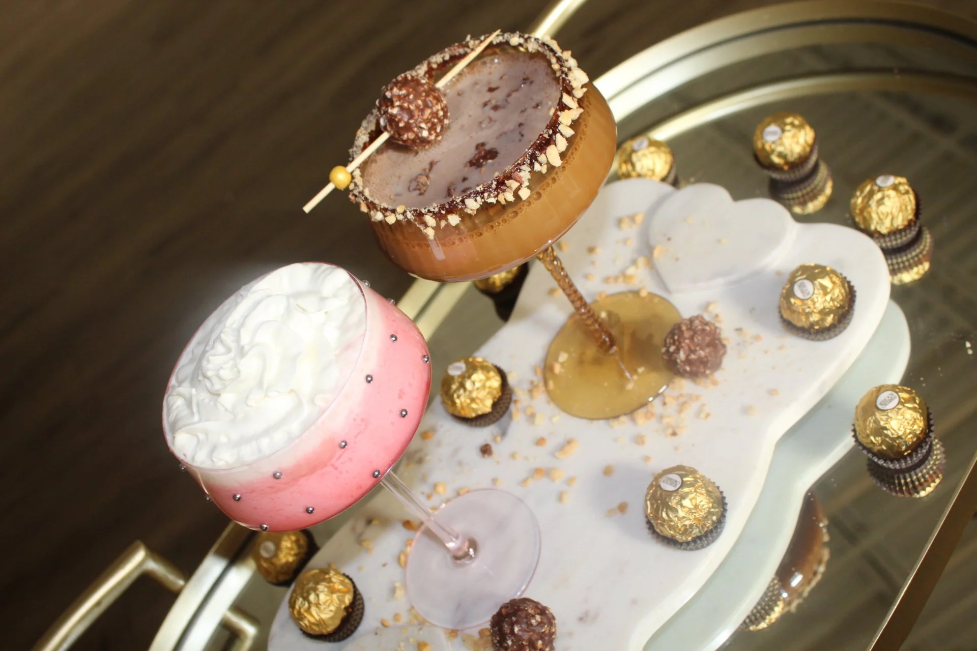 Elegant dessert platter with two colorful drinks, one pink with whipped cream and small silver decorative beads, and one brown with a salted caramel rim, served on a white tray surrounded by gold foil chocolates.