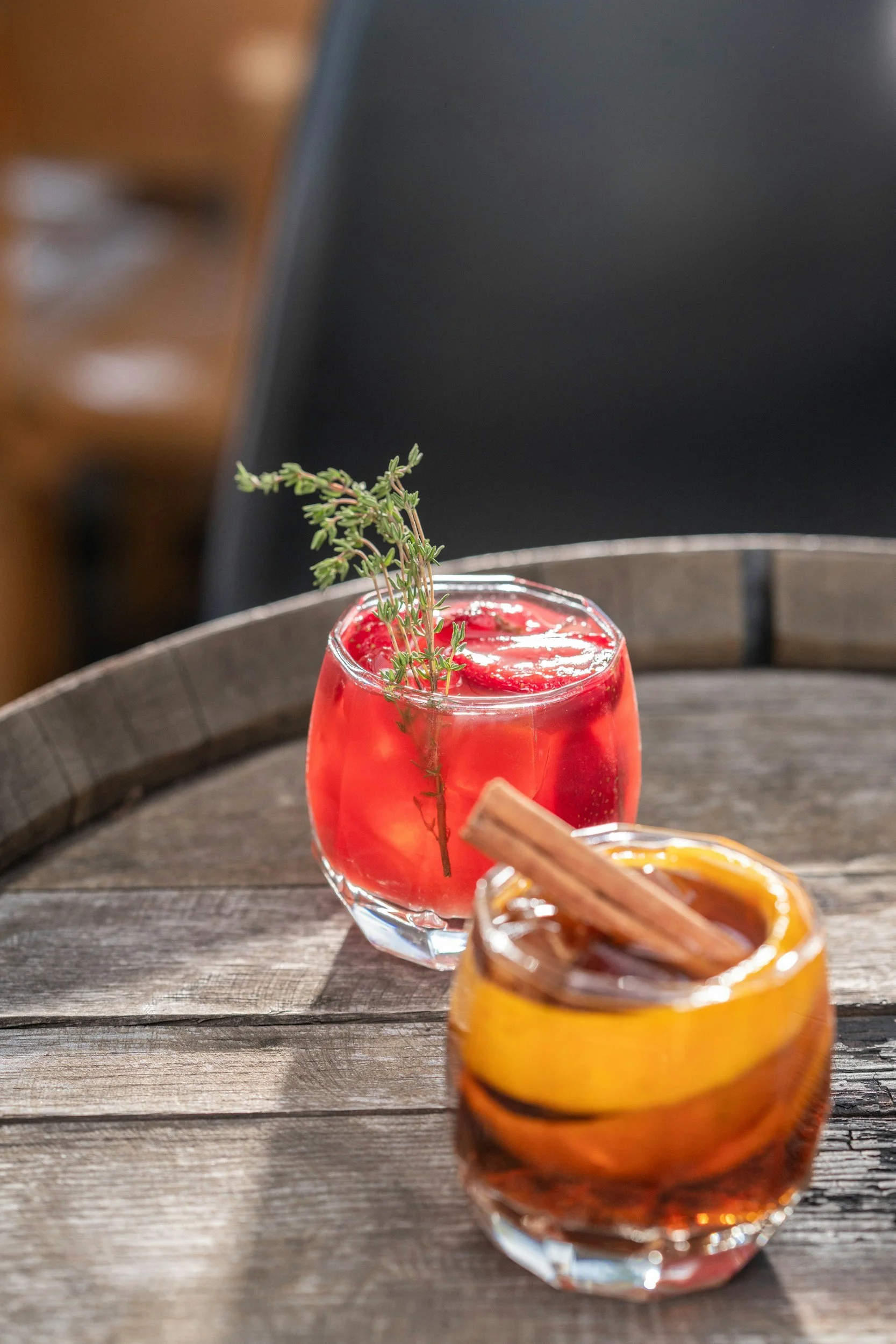 Two cocktails on a wooden table, one red with ice and a sprig of herbs, the other amber with a cinnamon stick and apple slices.