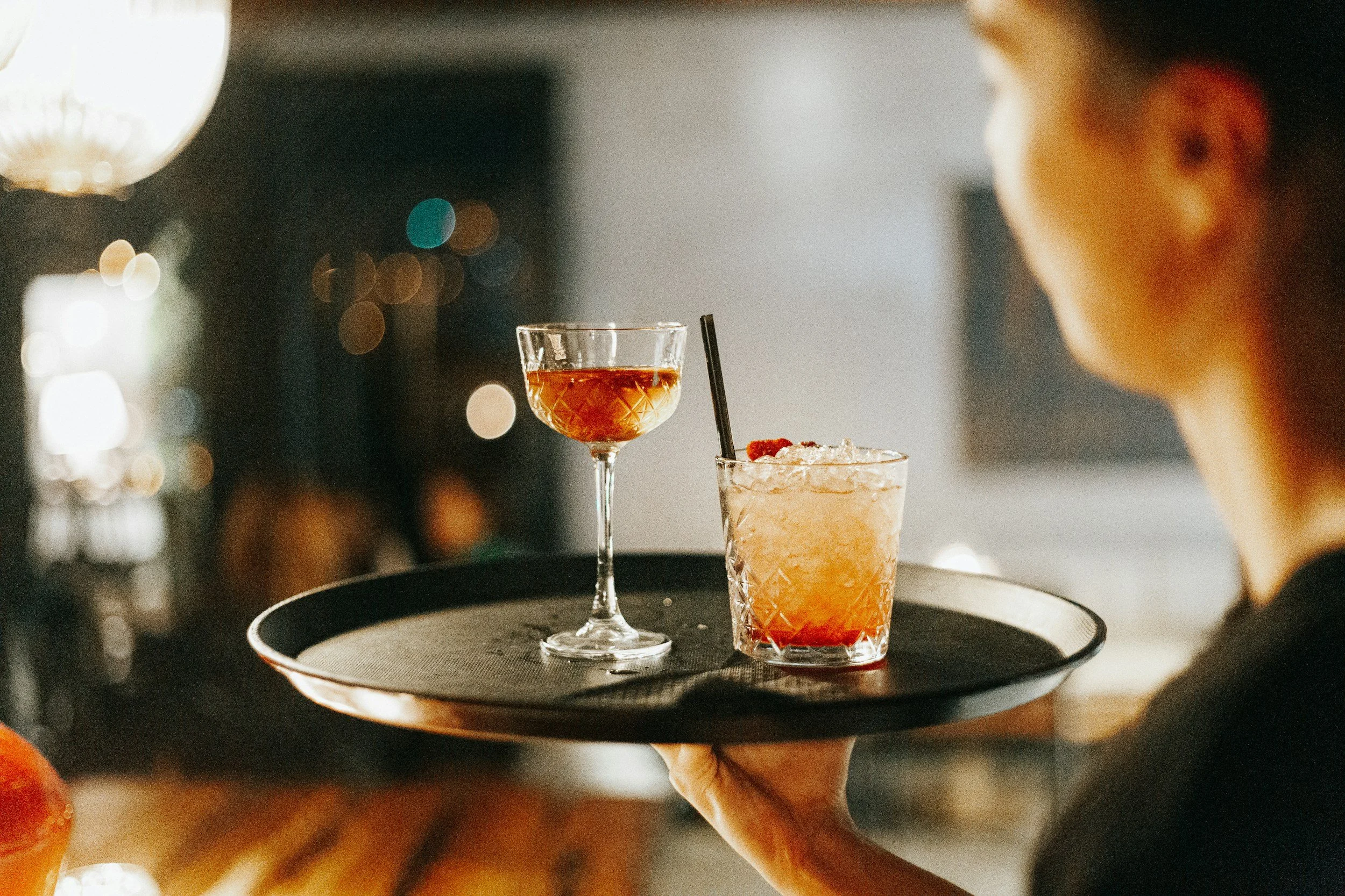A person holding a black tray with two cocktails on it, one in a tall glass and the other in a short glass with a straw, in a dimly lit bar or restaurant setting.
