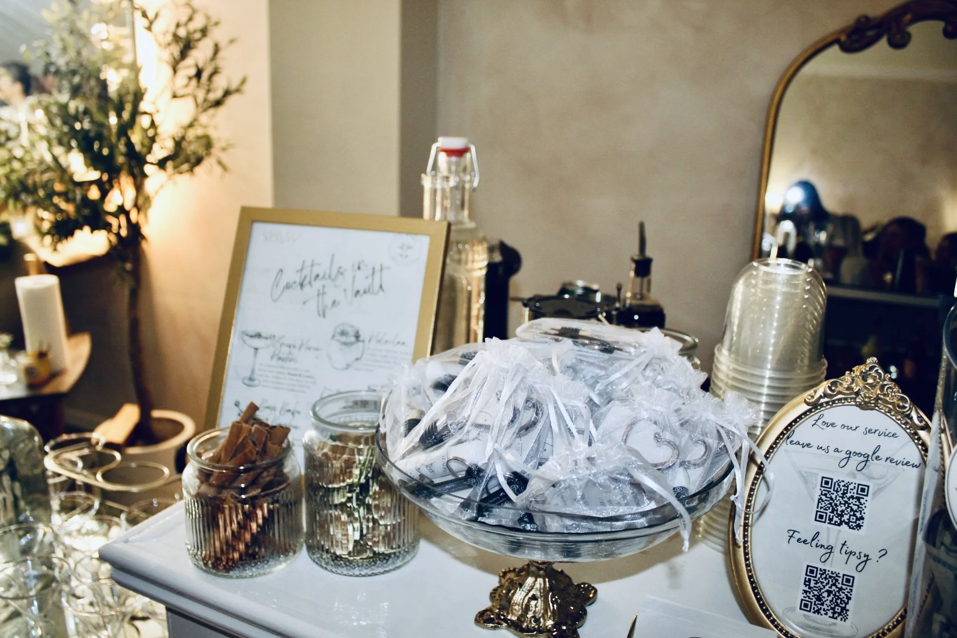 A table display of bar accessories with jars of cinnamon sticks, plastic stirrers, a framed drink menu, and QR codes for reviews at an event or bar setting.