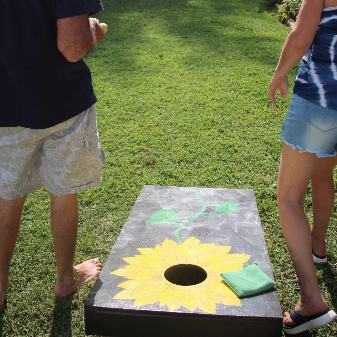 Two people playing the bean bag toss game outdoors on a grassy area, with a painted sunflower on the game board and a green bean bag nearby.