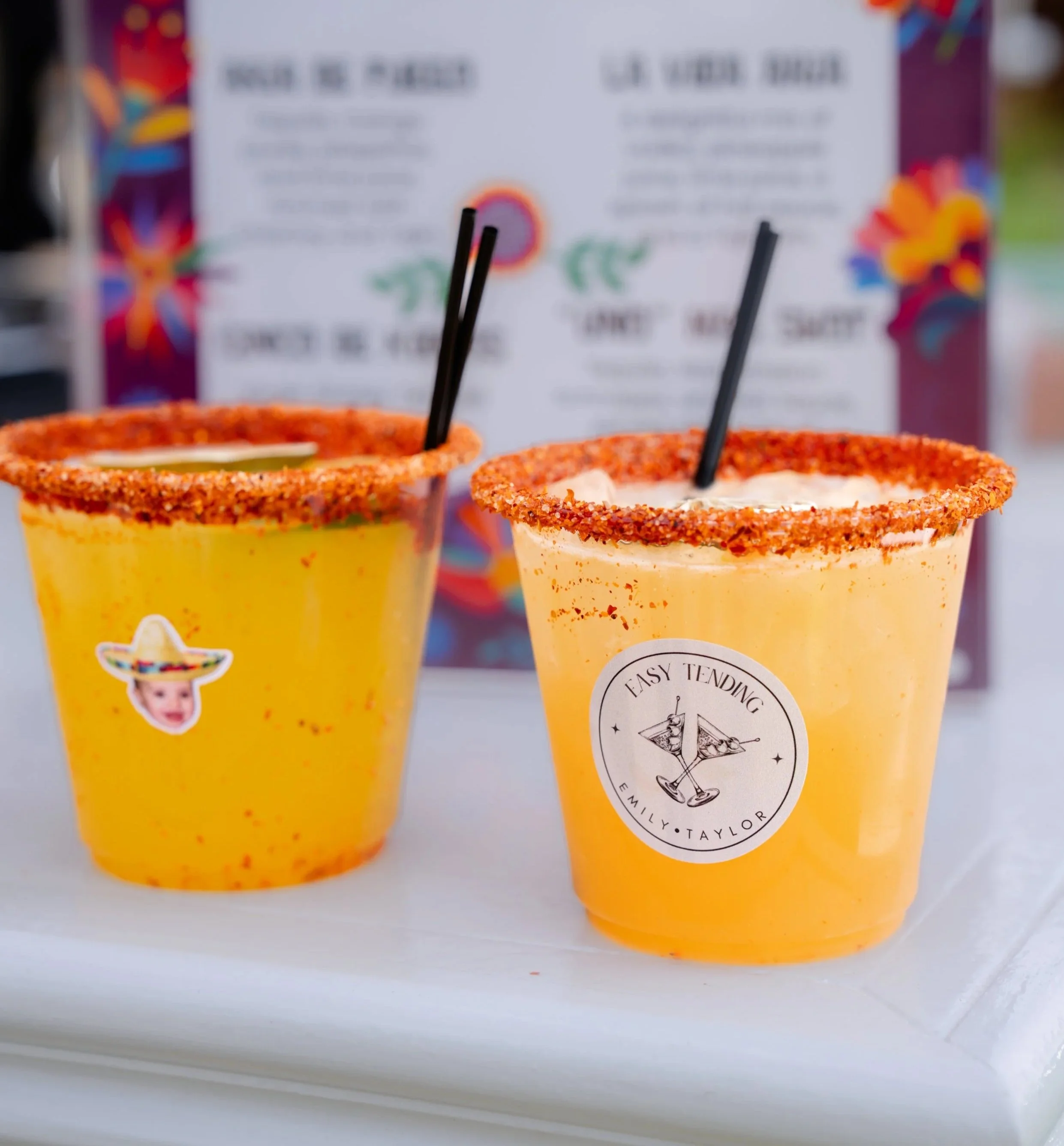 Two yellow frozen drinks with salted rim, one with a sticker of a baby wearing a sombrero and the other with a logo that says Easy Tending Emily Taylor, on a white surface with blurred menu in background.