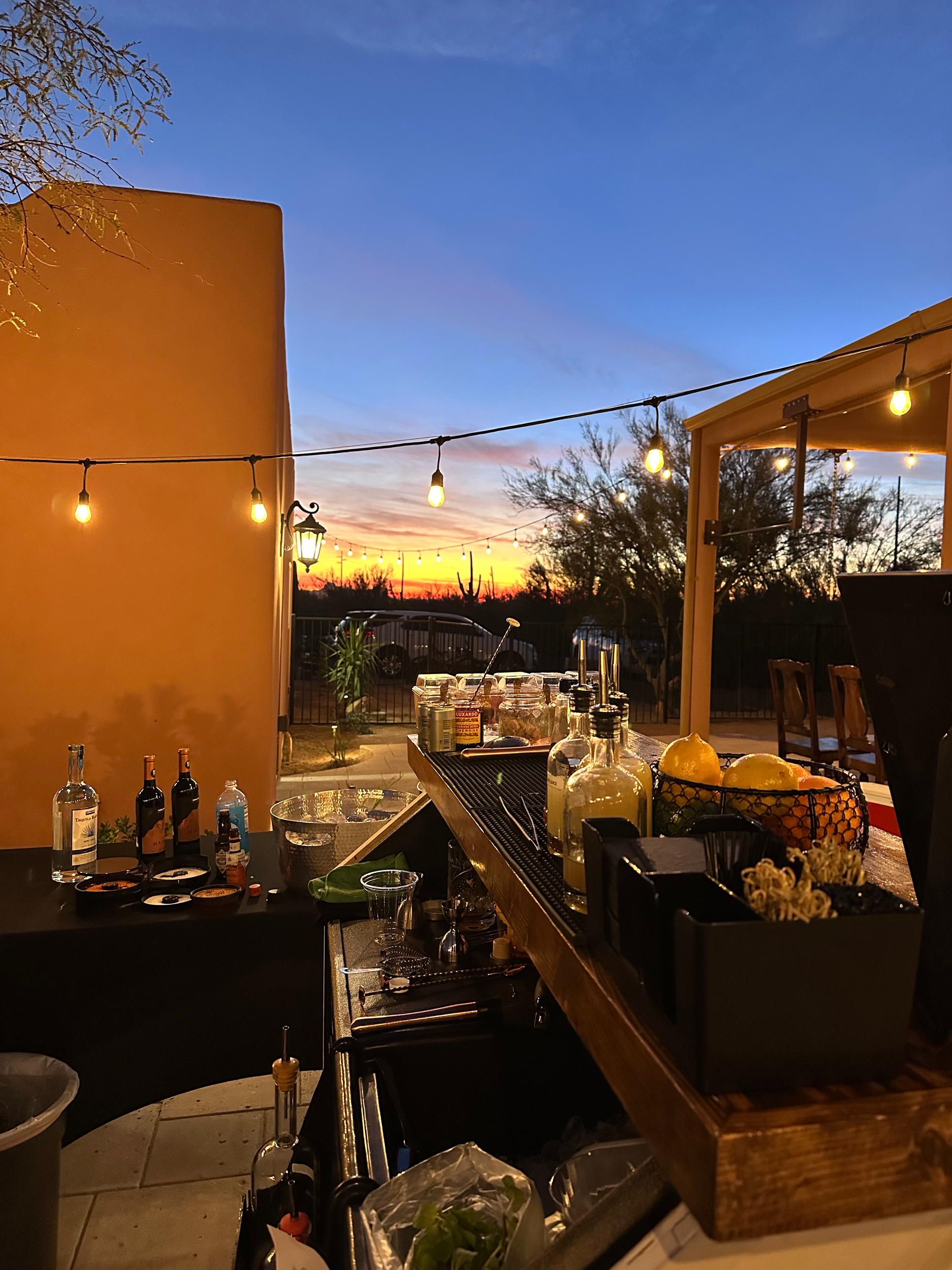 Outdoor bar setup on a patio at sunset with string lights, bottles of alcohol, glasses, lemons, and a view of the colorful sky.