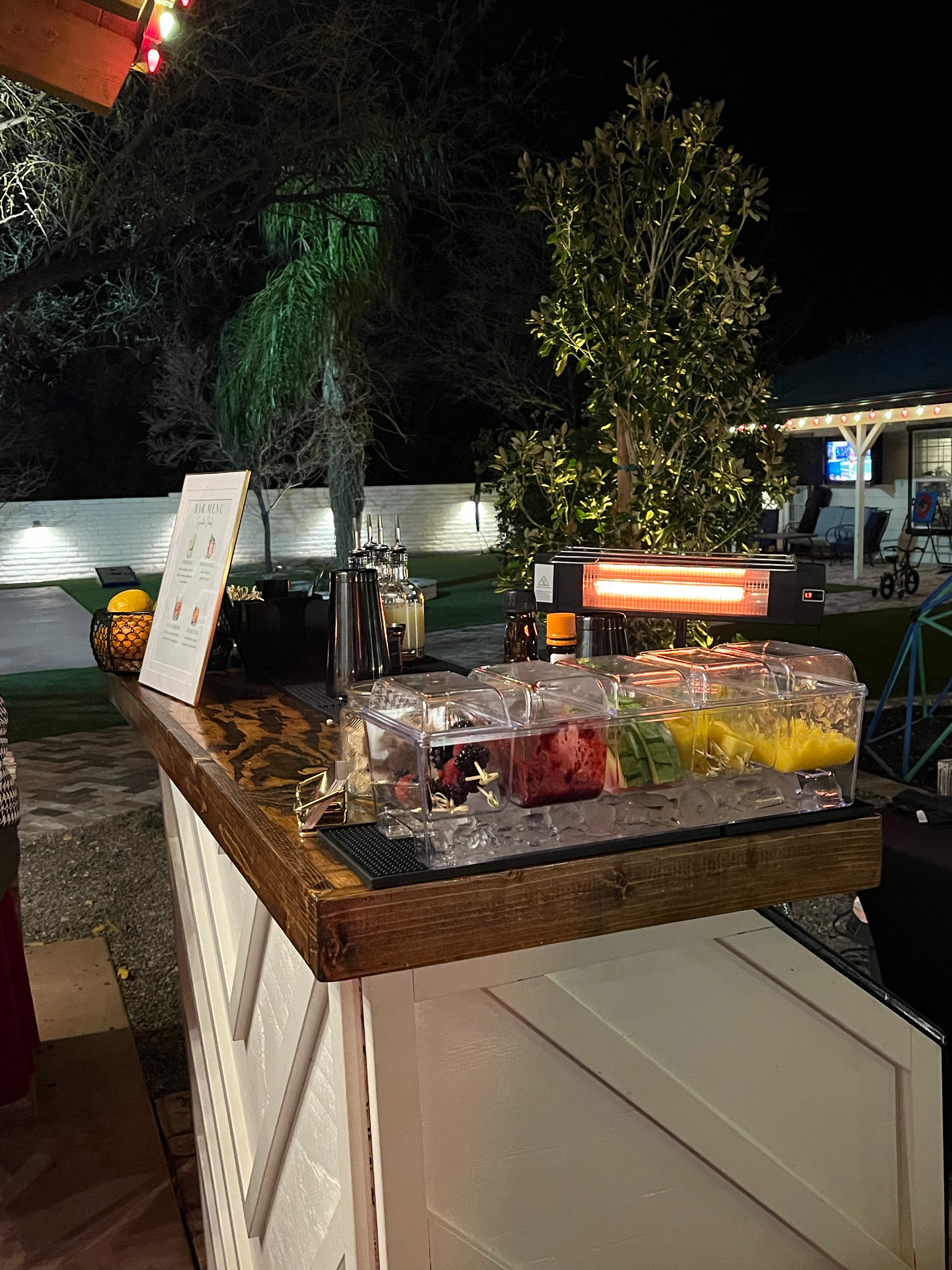 Outdoor beverage station with colorful fruit-infused water jugs, an electronic heater, and a menu sign, set against trees illuminated at night.