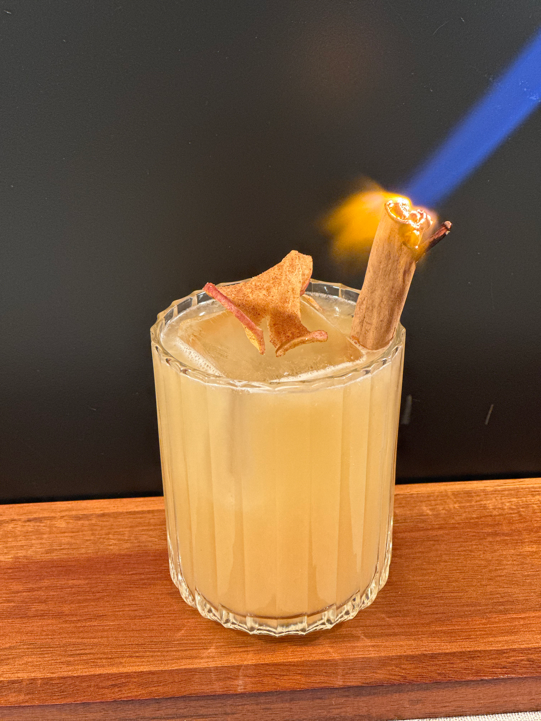 A glass of creamy yellow cocktail with a cinnamon stick and a star-shaped ginger cookie garnish, set on a wooden surface with a black background.