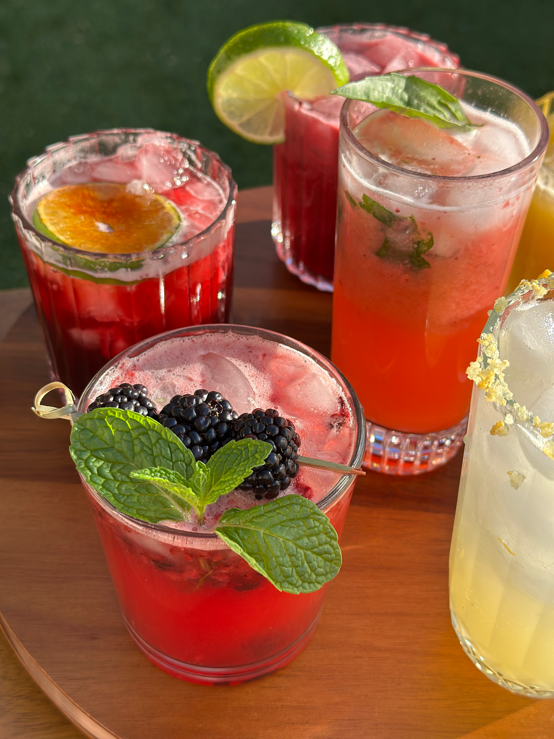 Four colorful cocktails on a wooden surface, garnished with lime slices, blackberries, and mint leaves.