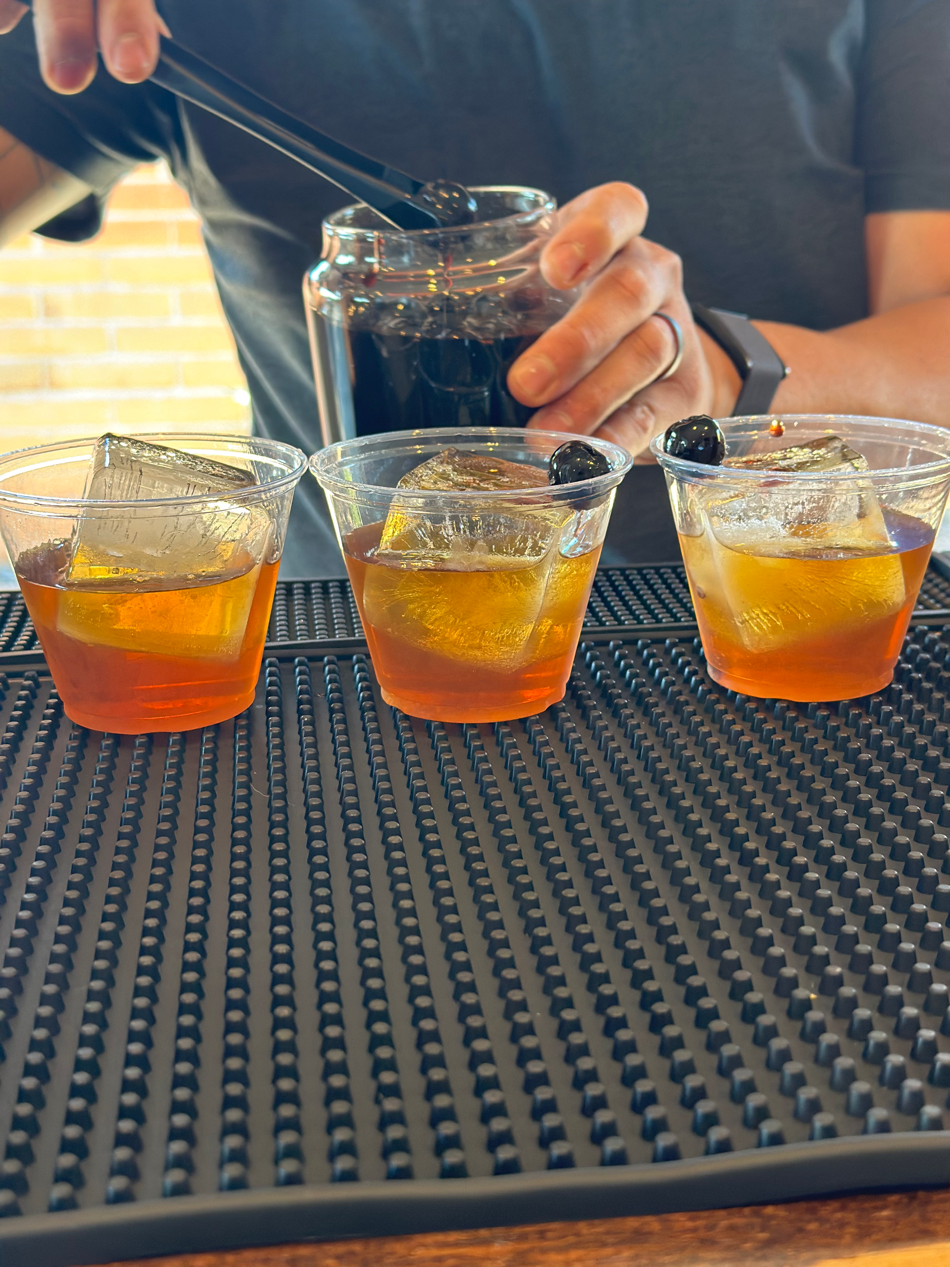 Three glasses of iced tea with lemon slices and ice cubes on a black rubber mat on a wooden table, with a person's hand holding a spoon.