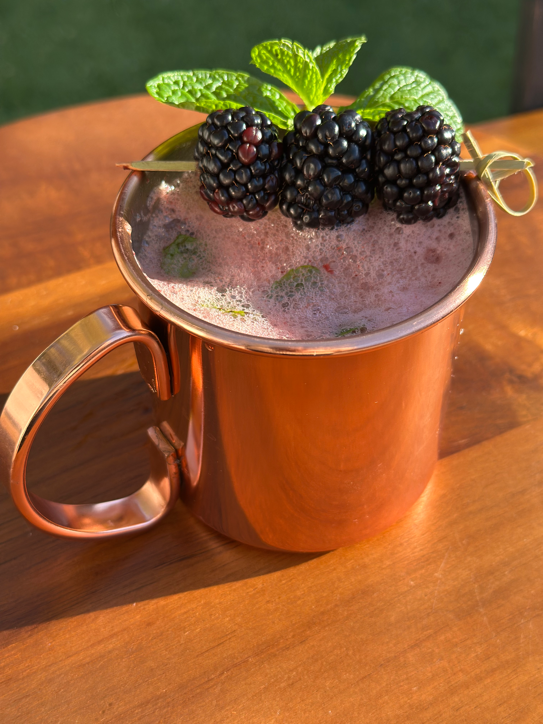 A copper mug filled with a blackberry cocktail garnished with fresh blackberries, mint leaves, and a bamboo skewer, sitting on a wooden surface.
