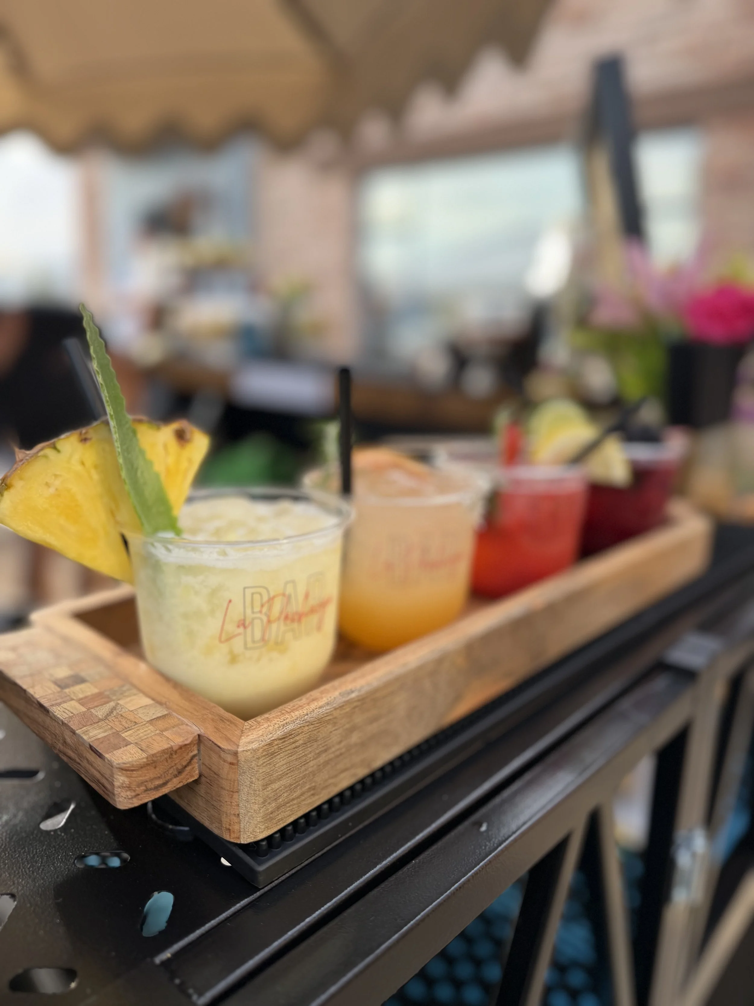Close-up of three colorful beverages in small glasses, garnished with pineapple and lime slices, displayed on a wooden tray at a bar or café.
