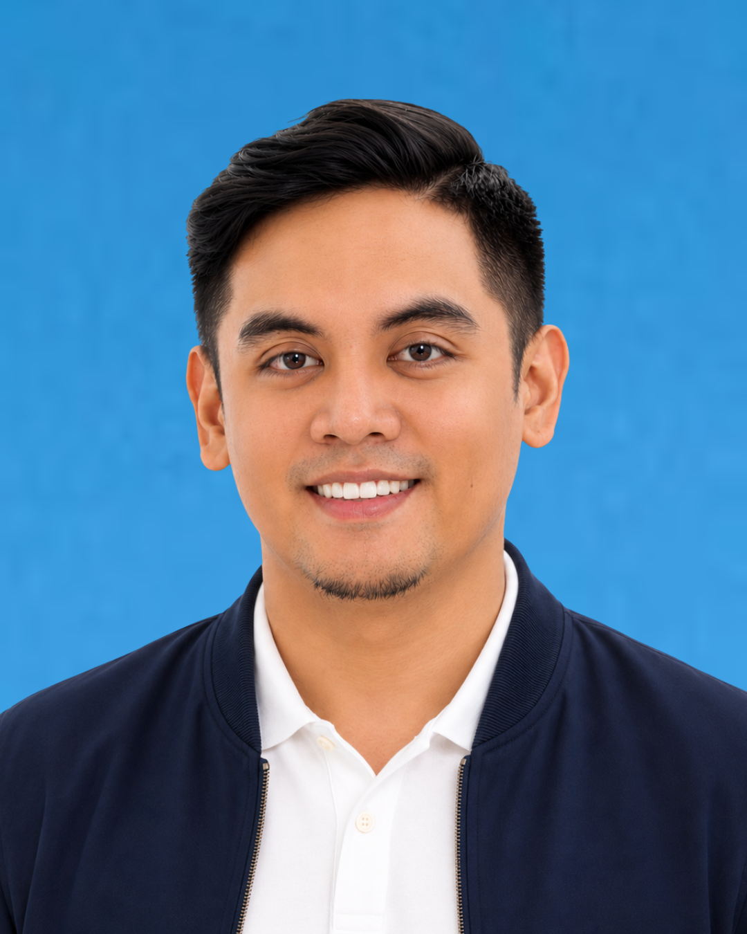 Professional headshot of a smiling man with dark hair, wearing a navy jacket and white collared shirt against a blue background.