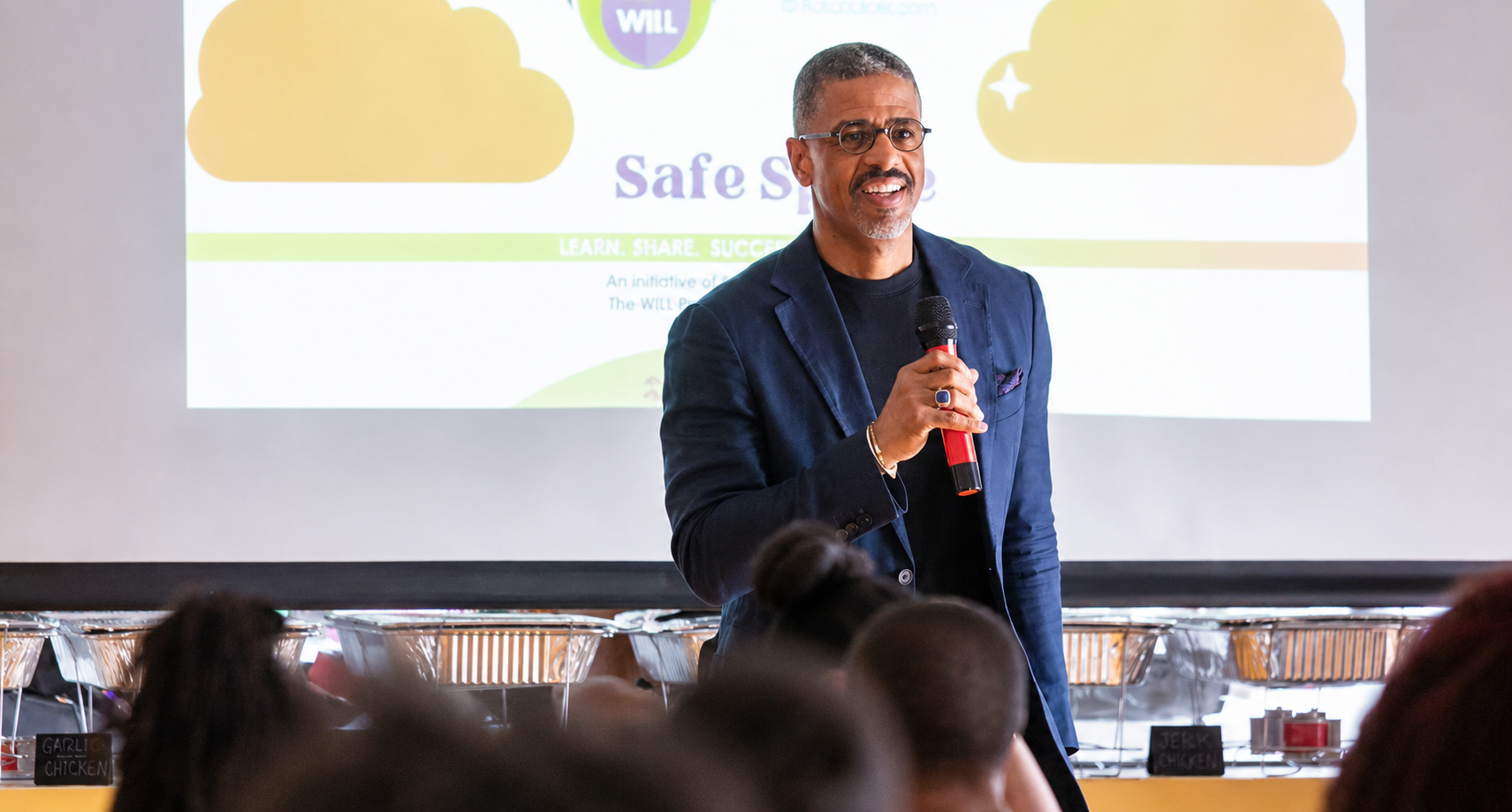 A man in a dark blazer holding a microphone stands in front of a presentation slide titled "Safe Space" during a talk or workshop. The background shows part of the slide with a cloud graphic and the phrase "LEARN, SHARE, SUCCEED." Audience members are visible in the foreground, and food labels like "GARLIC CHICKEN" and "JERK CHICKEN" are displayed on a buffet table behind the speaker.