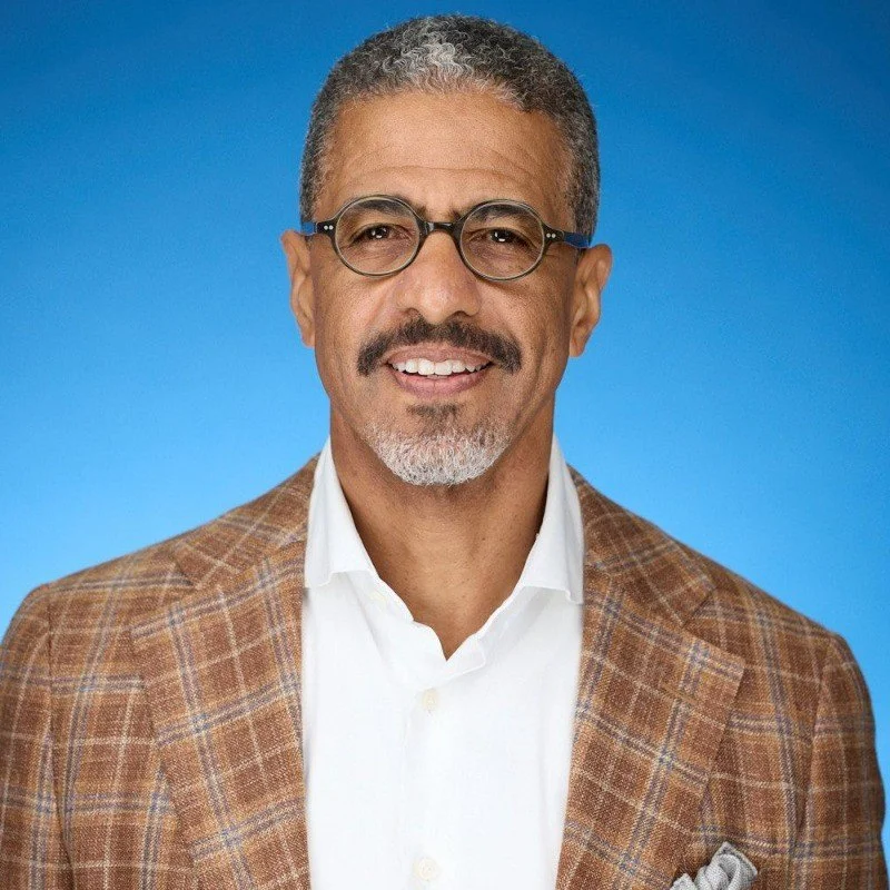 A middle-aged man with short gray hair and glasses, wearing a checkered brown blazer and white shirt, smiling against a blue background.