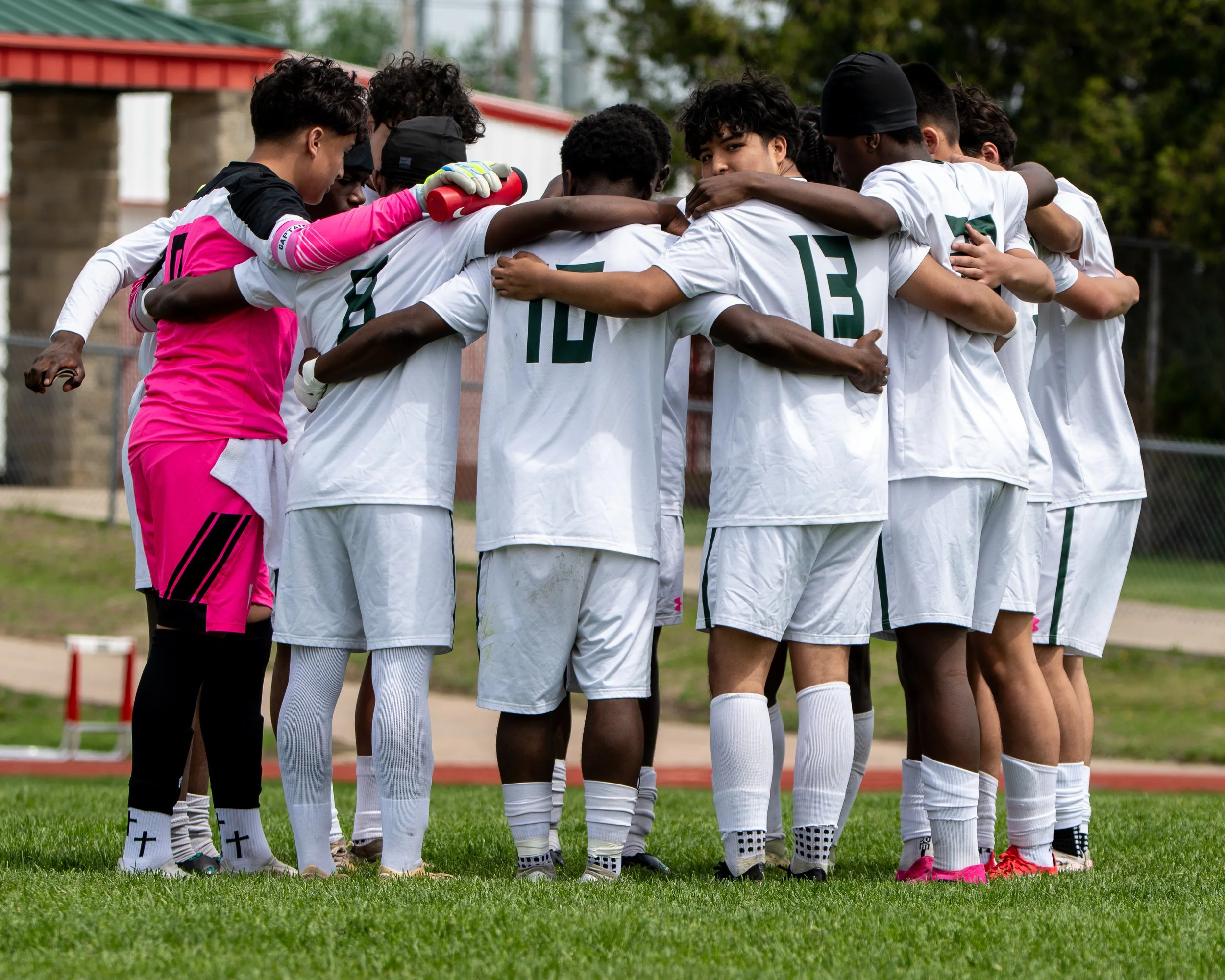 A group of young soccer players huddled together on the field, arms around each other, in a pre-game or motivational moment. The players are dressed in white jerseys, with one in pink goalie uniform, against a background of green trees and a red-brown fence.