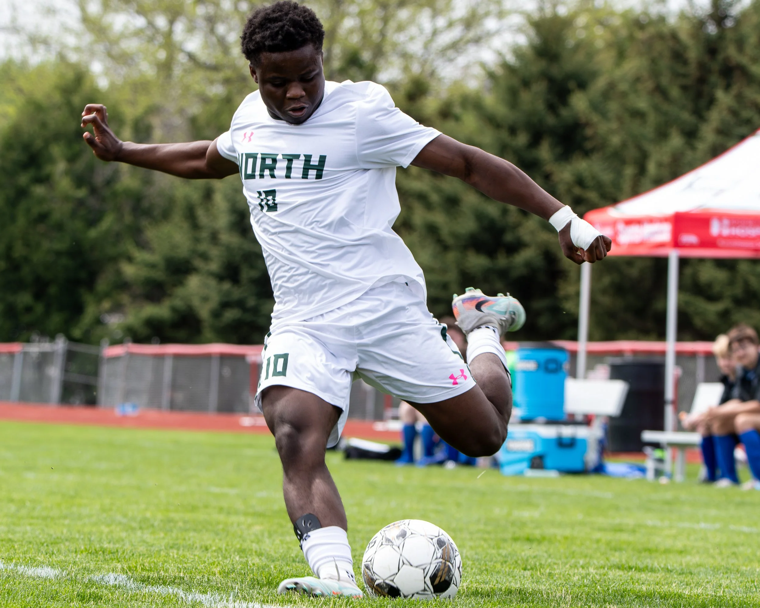 A young male soccer player with dark skin and curly hair wearing a white jersey with 'NORTH' printed on it, playing soccer outdoors on a grassy field during daytime. He is kicking a black and white soccer ball, with a focused expression. In the background, there are trees and a red tent.