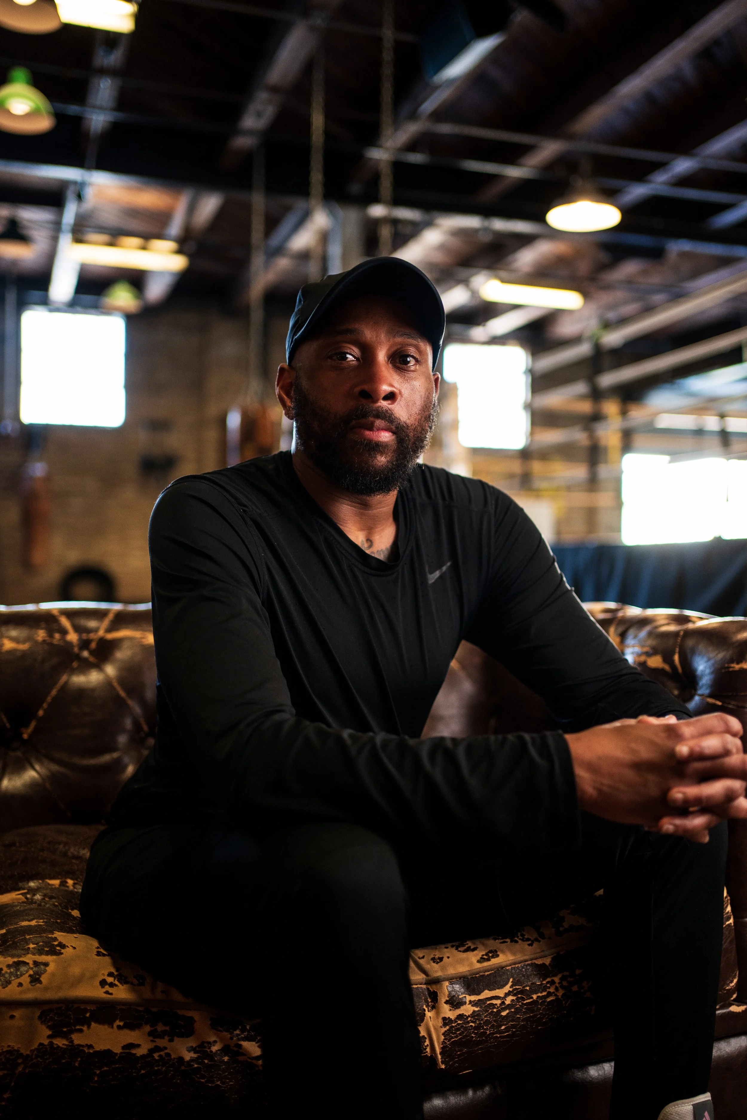 An African American man sat on a brown leather coach in front of a boxi g ring in a boxing gym,  he's wearing a black cap and a black long sleeve sports top