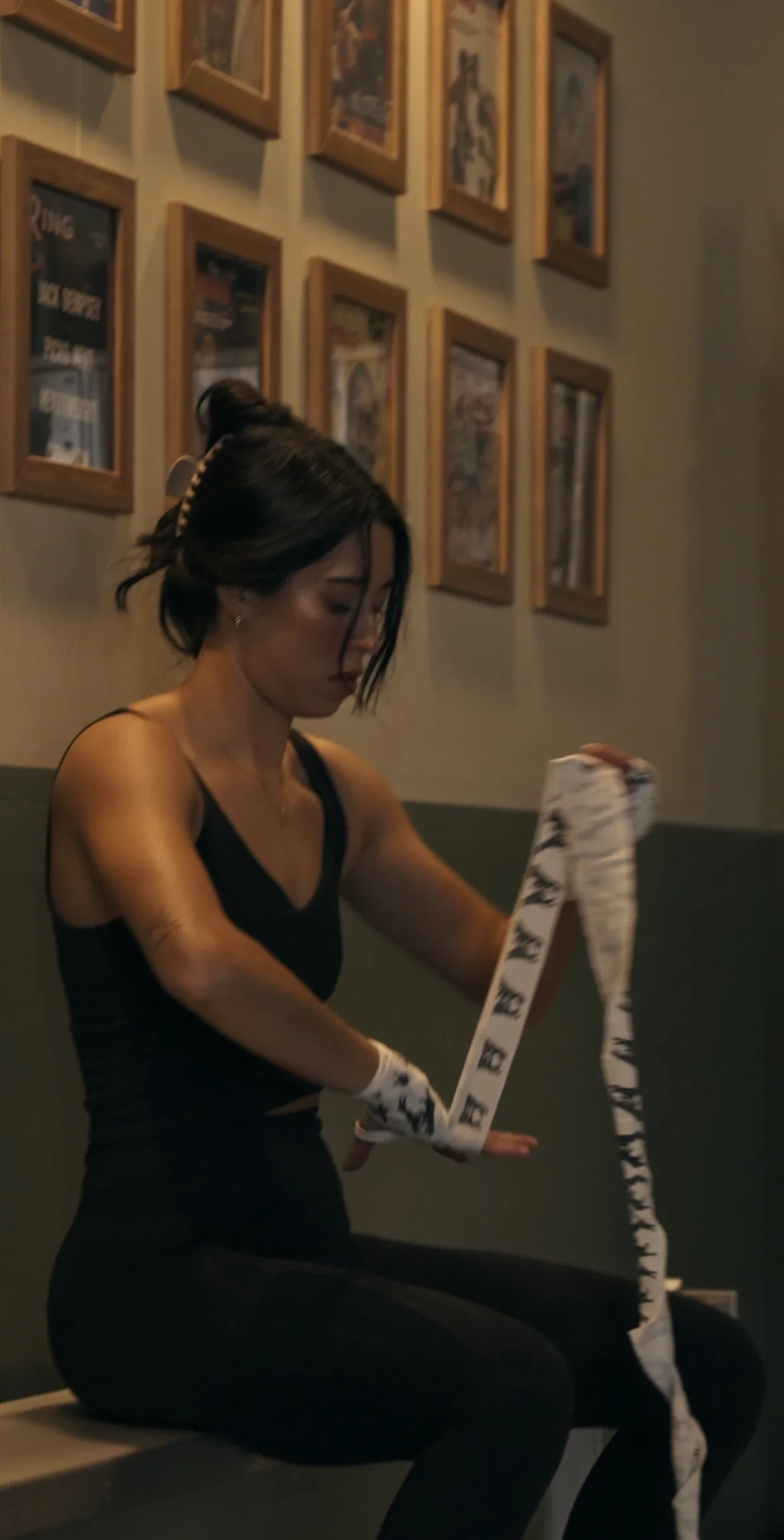 A lady sitting on the bleachers in a boxing gym with hair fallen down her face while she puts on her boxing hand wraps.