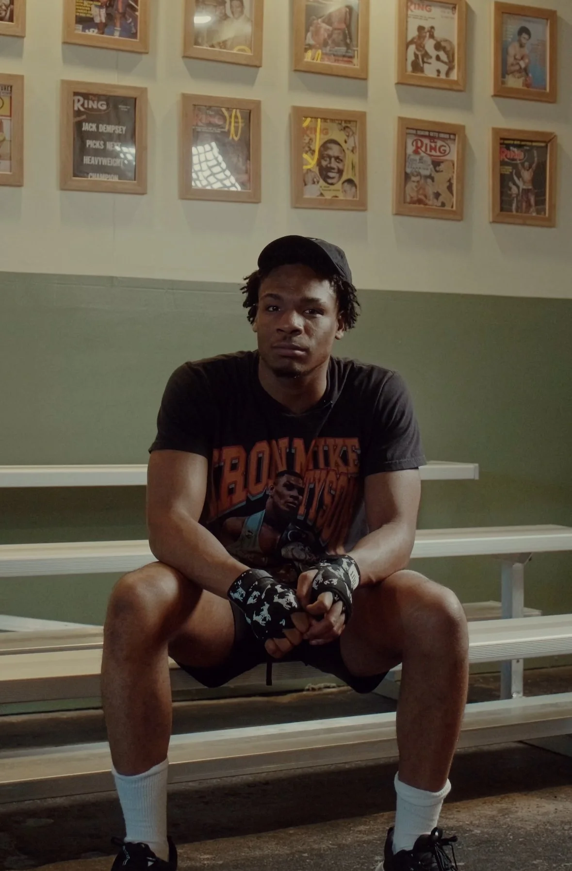 African American man wearing a black t shirt and black hat sat on a bleacher in a boxing gym with boxing hand wraps on.