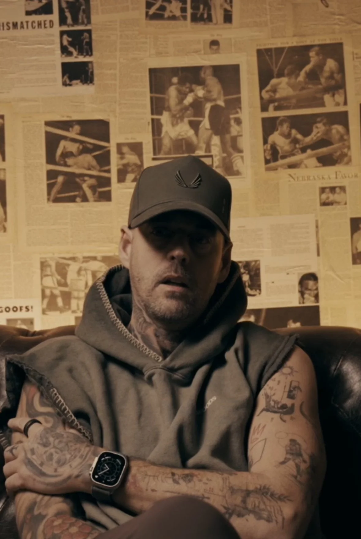 Gentlemen wearing a green hat and a sleeveless hoodie with tattoos sitting on a coach in front of a wall with vintage boxing magazine pages.