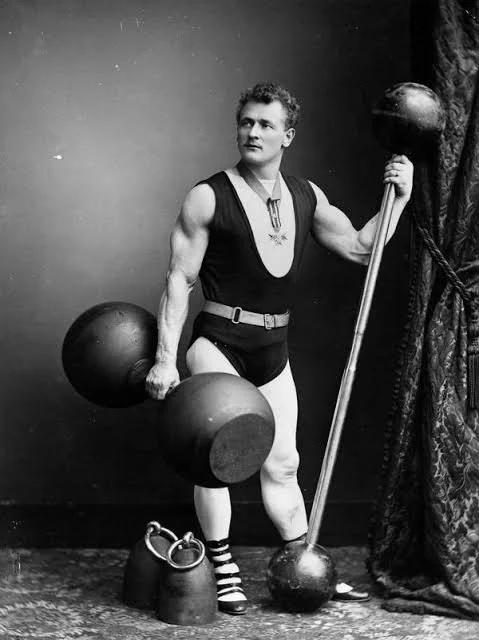 Black-and-white photo of a muscular man dressed in vintage circus or strongman attire, holding a large dumbbell in one hand and a pole in the other, standing next to a curtain.