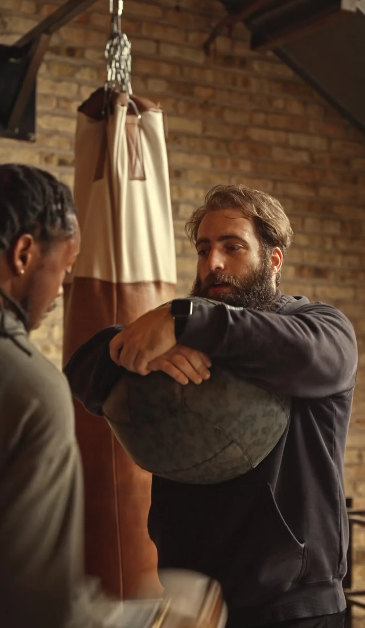 two men discussing boxing techniques,  one man has a beard and is holding a large ball in front of a boxing heavy bag.