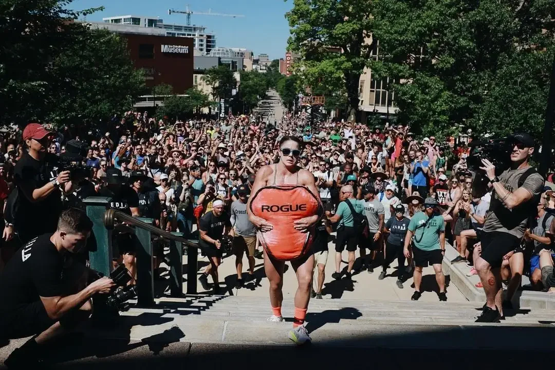 A woman participating in a fitness event, carrying a large orange Rogue-branded sandbag, surrounded by a crowd of spectators and cameras on stairs outdoors.