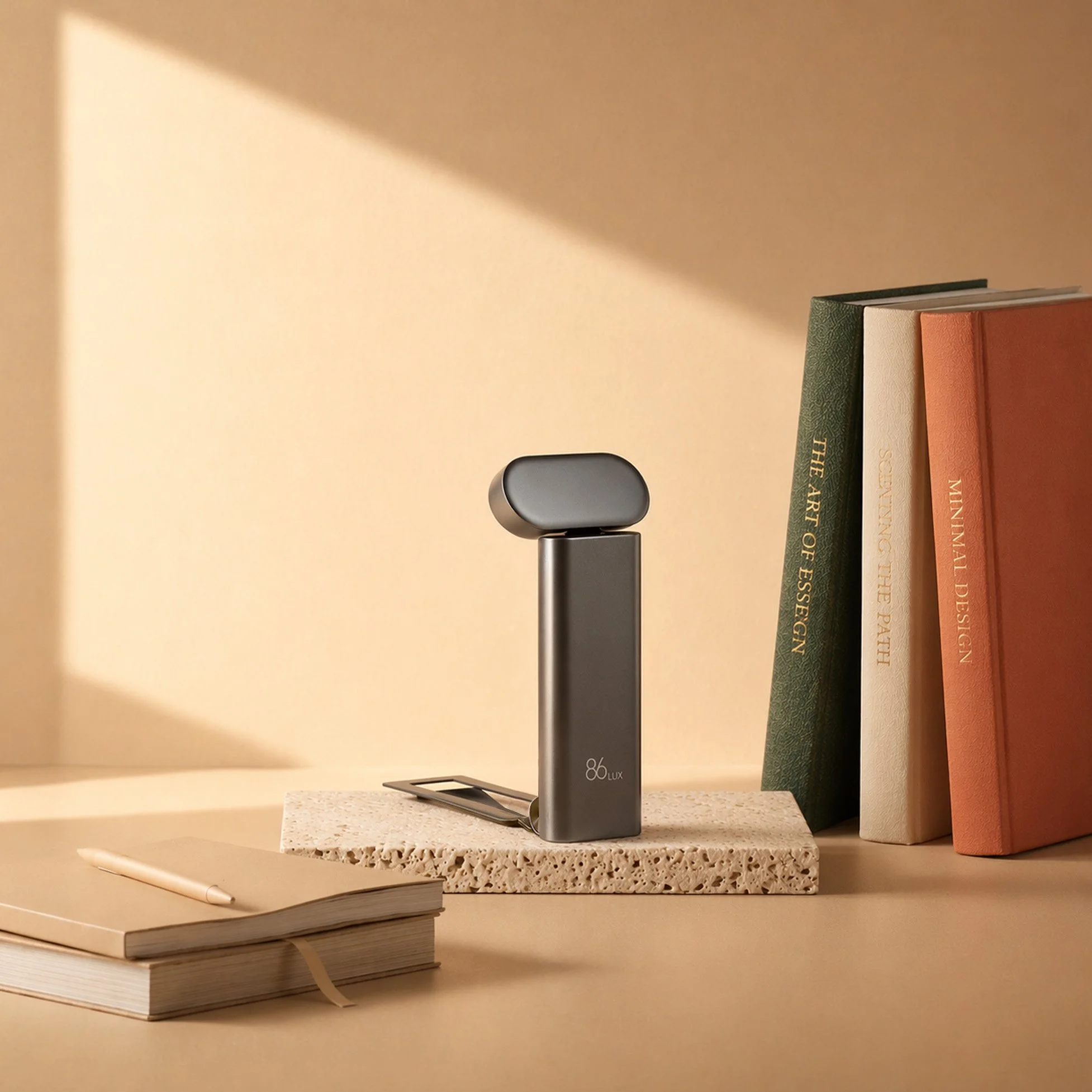 A desk setup with a tablet, a closed notebook, a pen, and a stack of books. The background has a beige wall with a section of shadow.