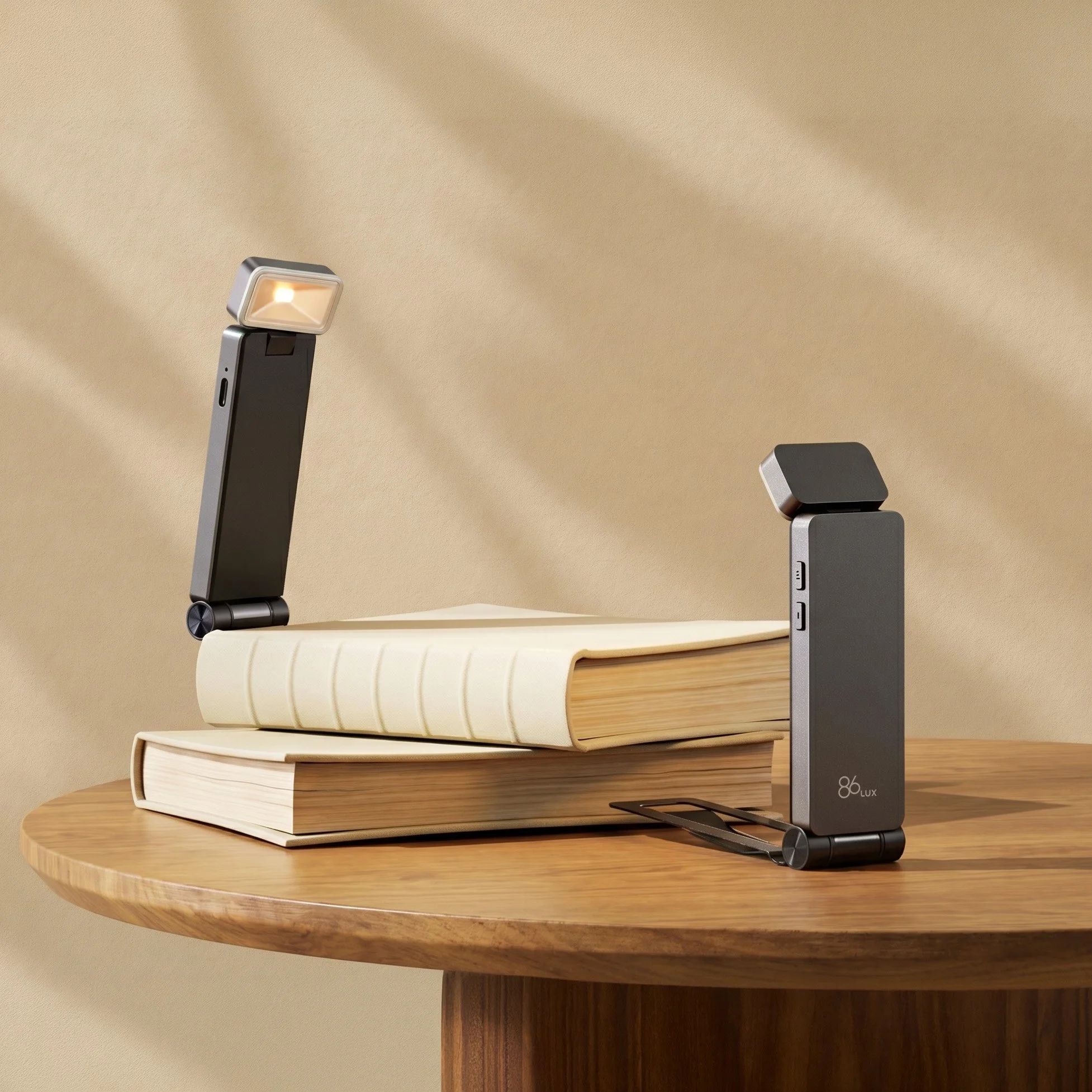 Close-up of a modern desk lamp with a black and gray design illuminating a stack of three closed books on a round wooden table against a beige wall.