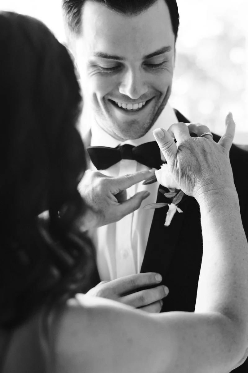 Groom smiling as his bow tie and boutonniere are adjusted during wedding preparations, captured in black and white.