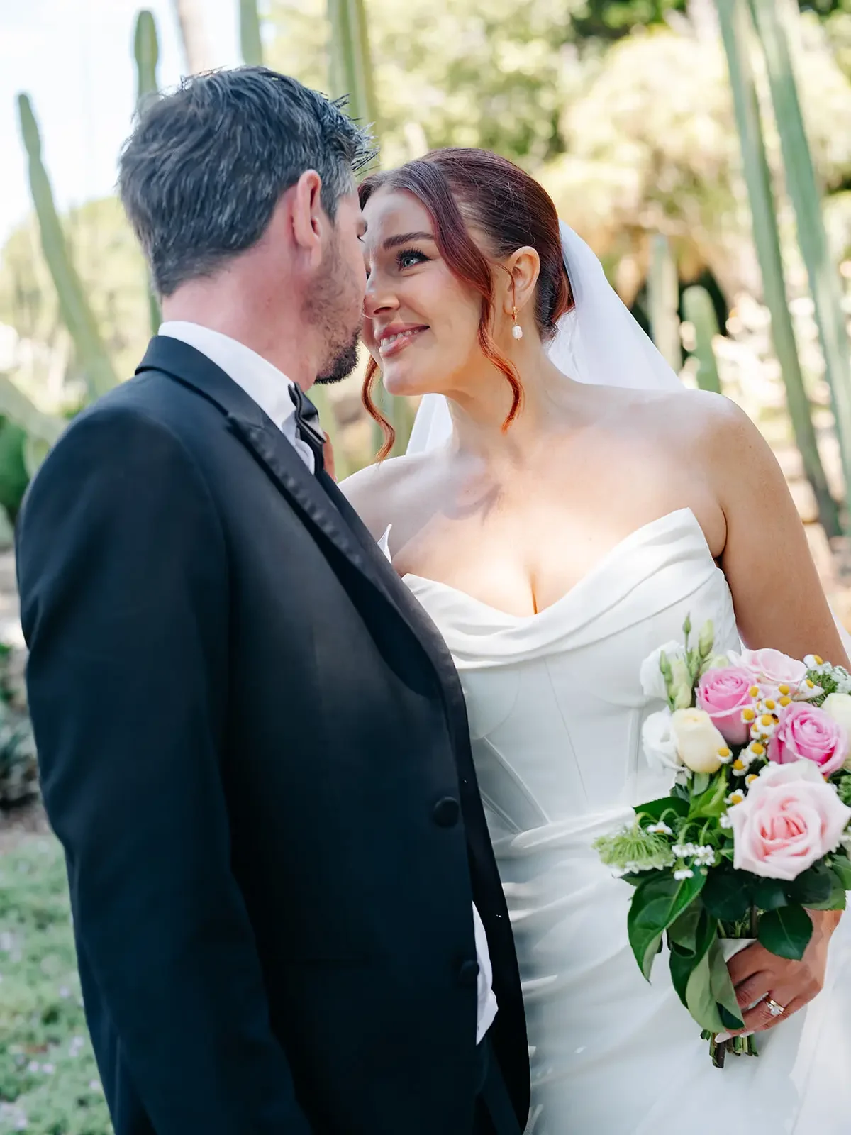 Bride holding a pink and white rose bouquet while smiling at her groom during an outdoor wedding portrait session.