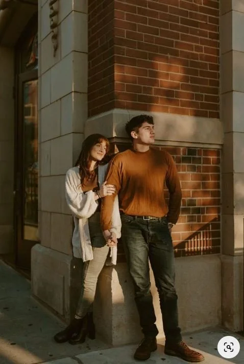 A young woman and man stand together under a brick and stone building at sunset, with the woman resting her arm on the man's shoulder.