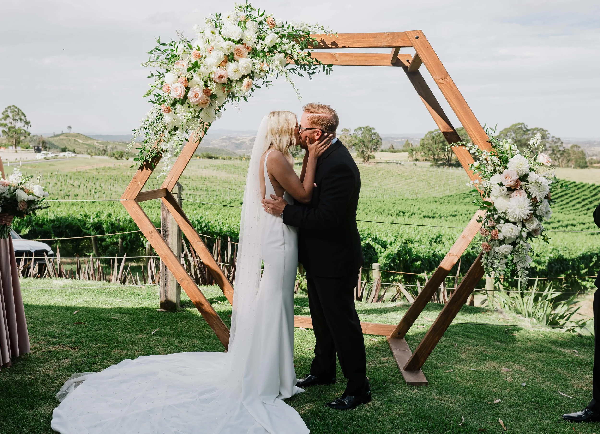 Bride and groom kissing beneath a floral wooden wedding arch overlooking a scenic countryside vineyard.