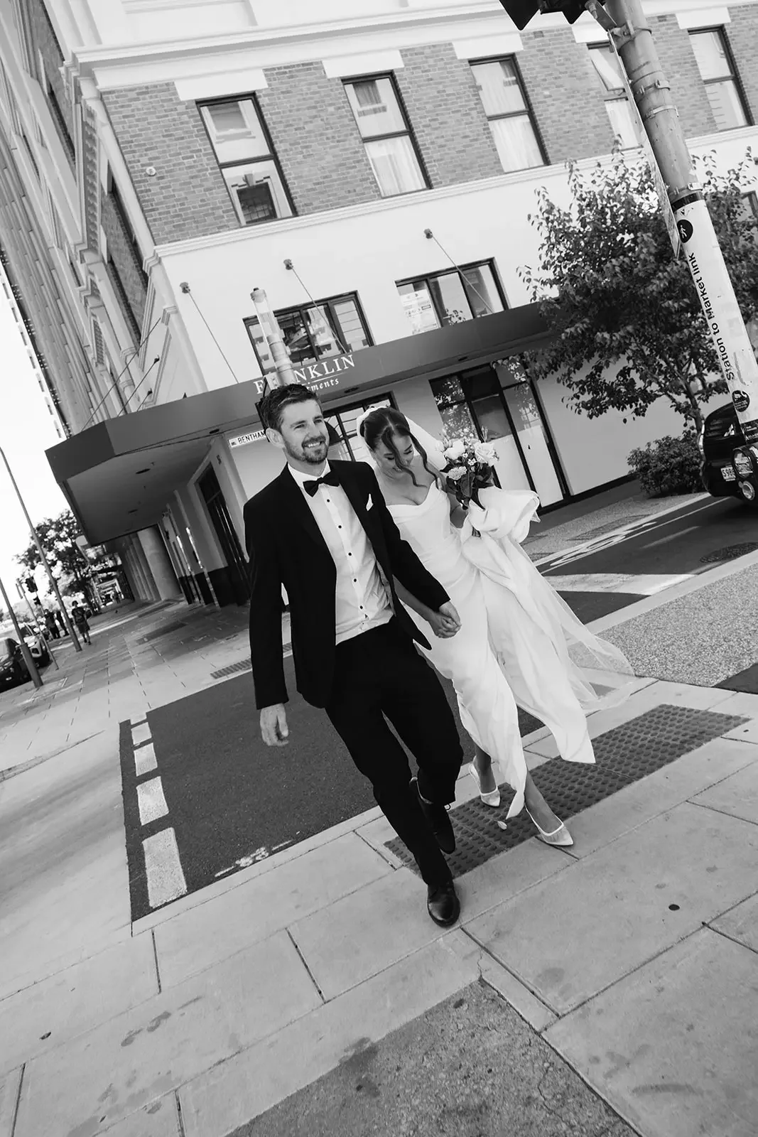 Black and white candid wedding photograph of a bride and groom walking hand in hand through a city street.