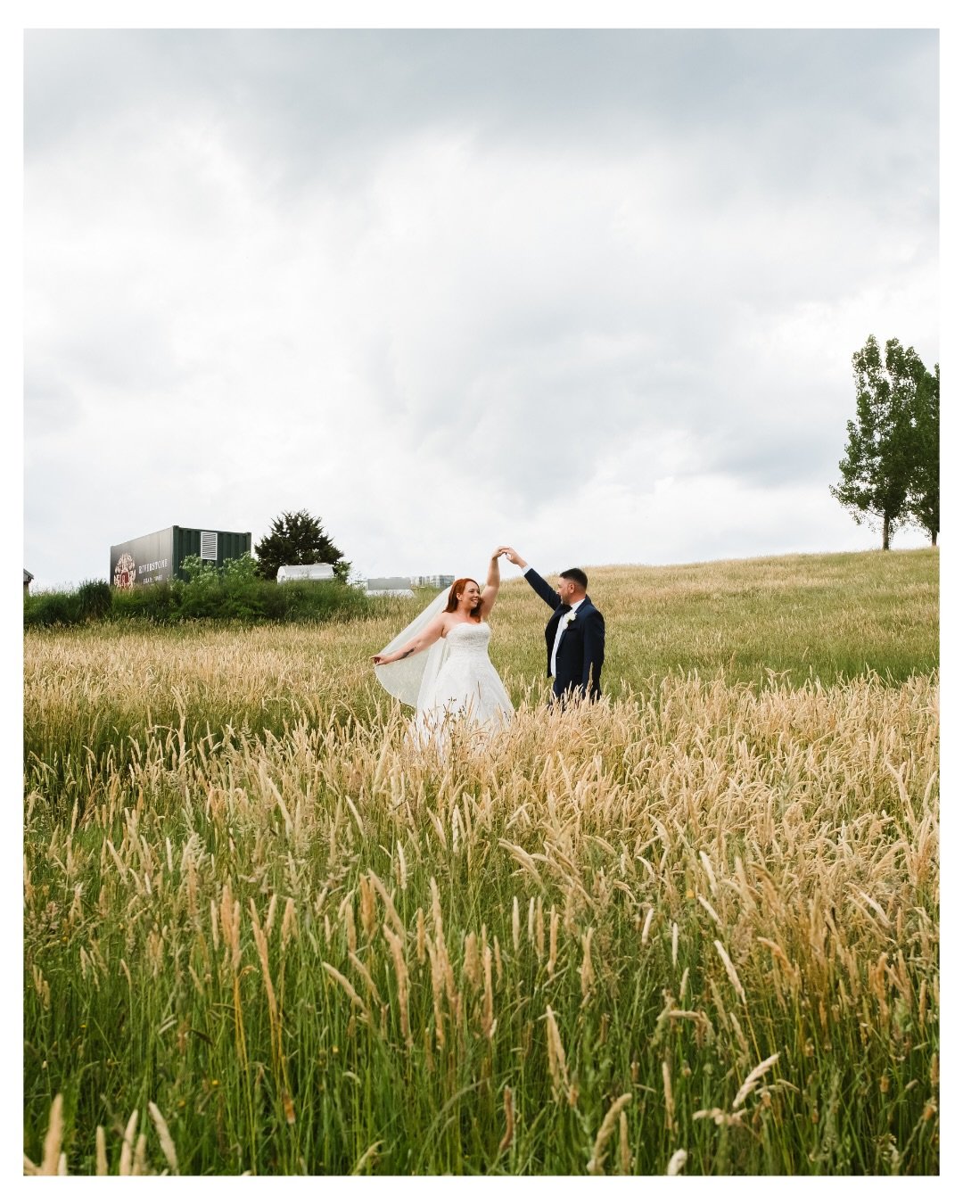 Bride and groom dancing in a grassy field beneath an overcast sky during their outdoor wedding photoshoot.