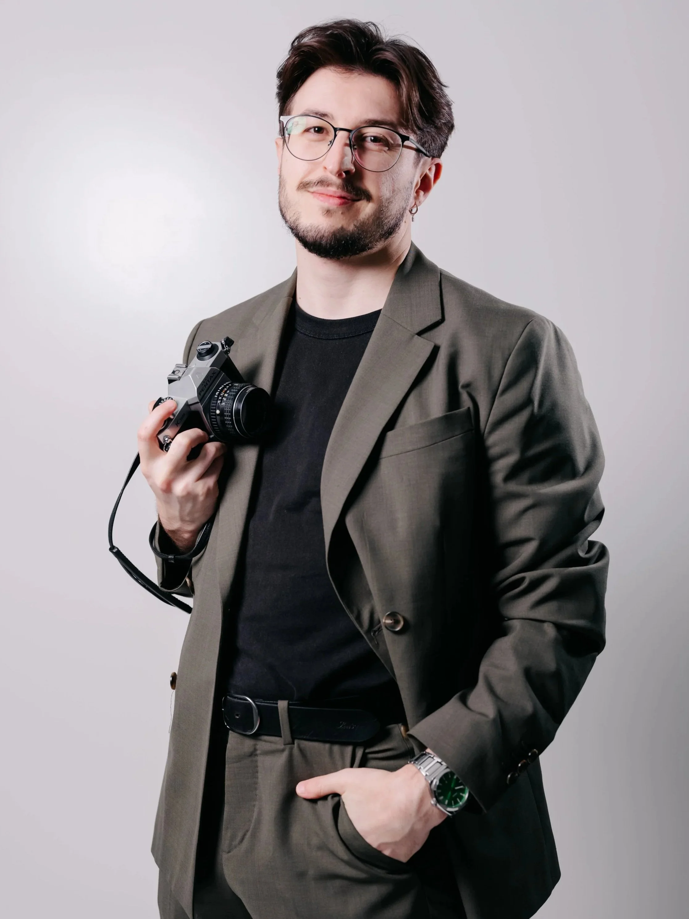 A young man with glasses and a beard, wearing a black denim jacket, holding a camera, standing against a dark background.