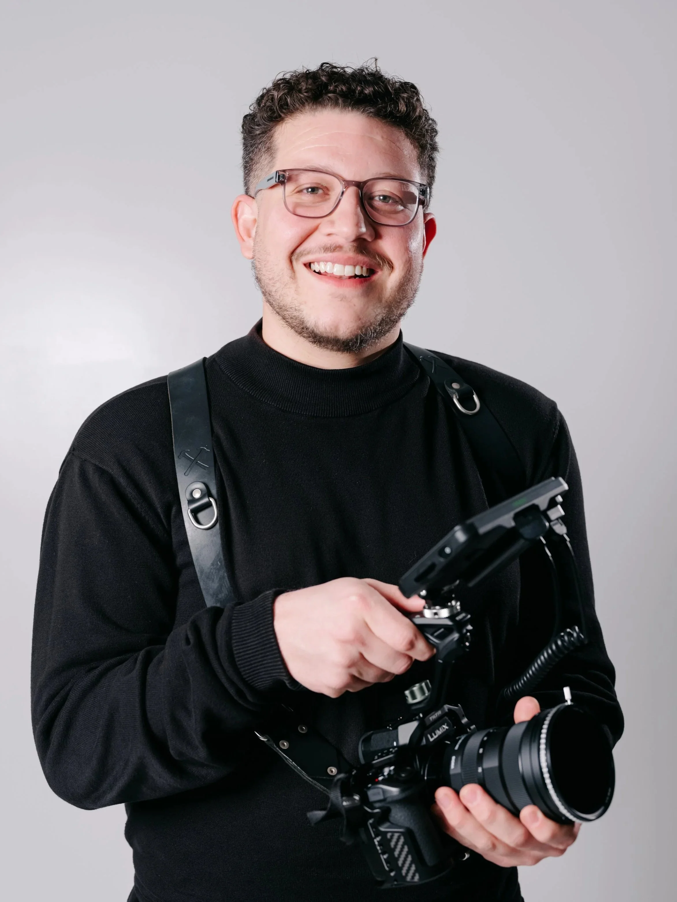 Smiling man with glasses holding a camera with a mounted accessory, standing against a dark background.