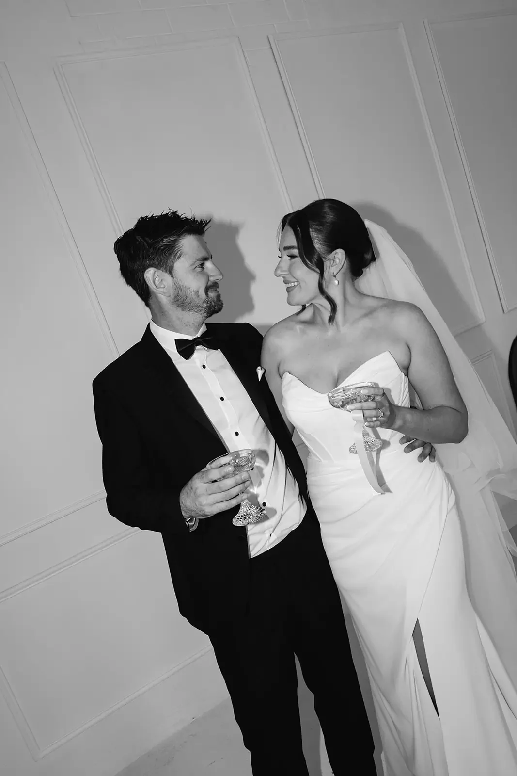 Black and white wedding portrait of a bride and groom smiling at each other while holding champagne glasses.