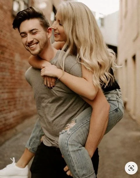 A young man giving a piggyback ride to a young woman on a city street, both smiling and enjoying the moment.