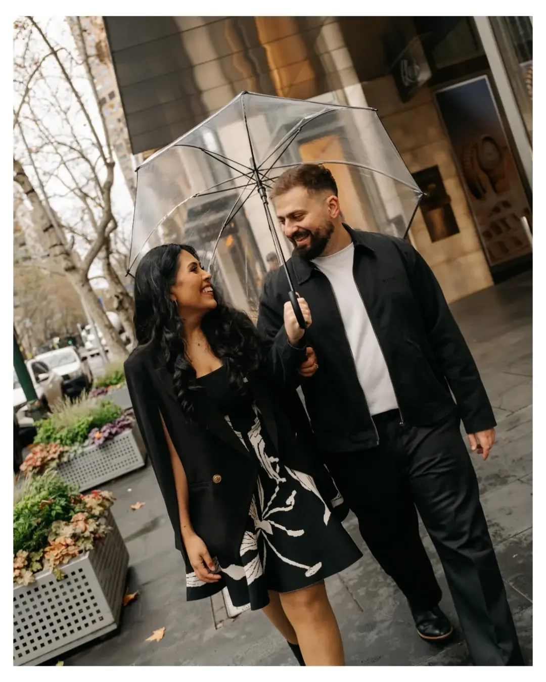 Couple smiling beneath a clear umbrella on a city street during a relaxed rainy-day engagement photography session.