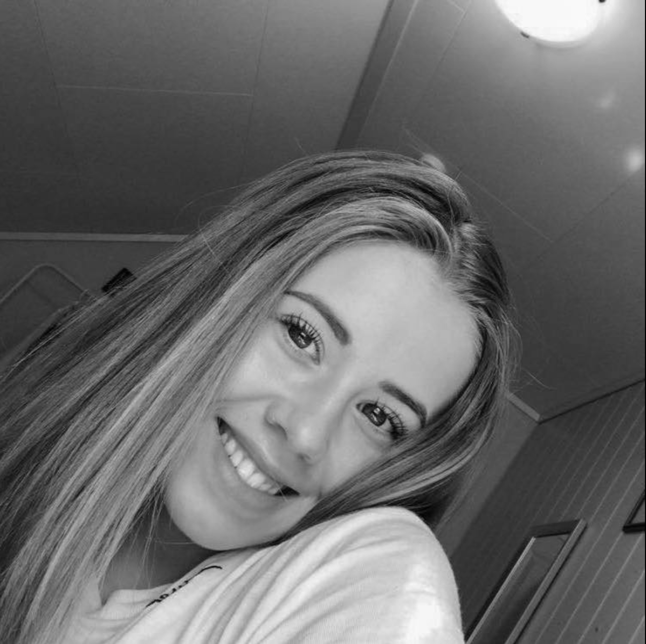 Et svart-hvitt bilde av en smilende kvinne med langt, rett hår, som tar en selfie i innendørs omgivelser.